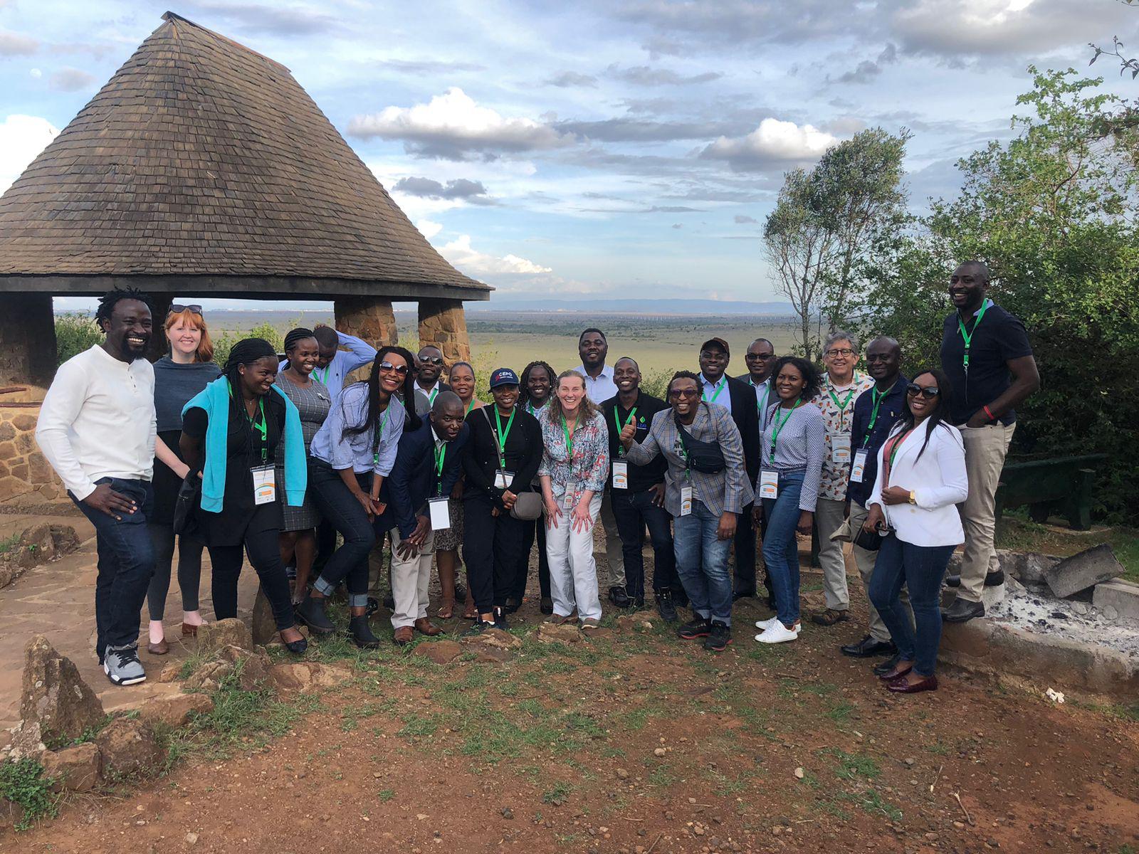 A group of smiling participants posing together at Nairobi National Park in front of a walkway, hut, trees, and blue sky.